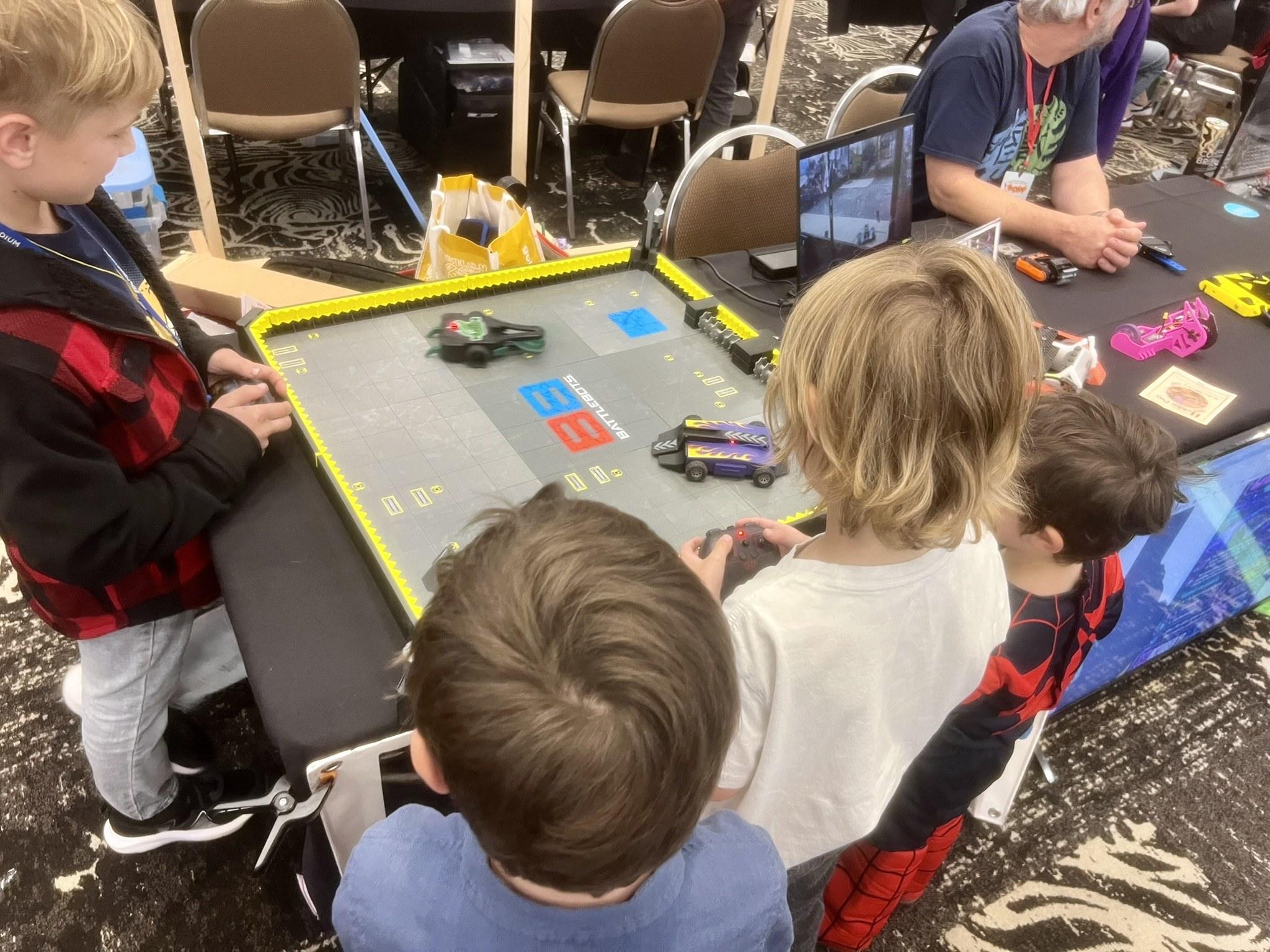 Kids gathered around a robot arena table, driving small robots with controllers.